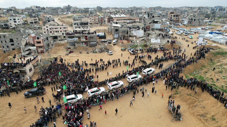 Aerial view of a large crowd gathered in a war-torn city, with Red Cross vehicles present.