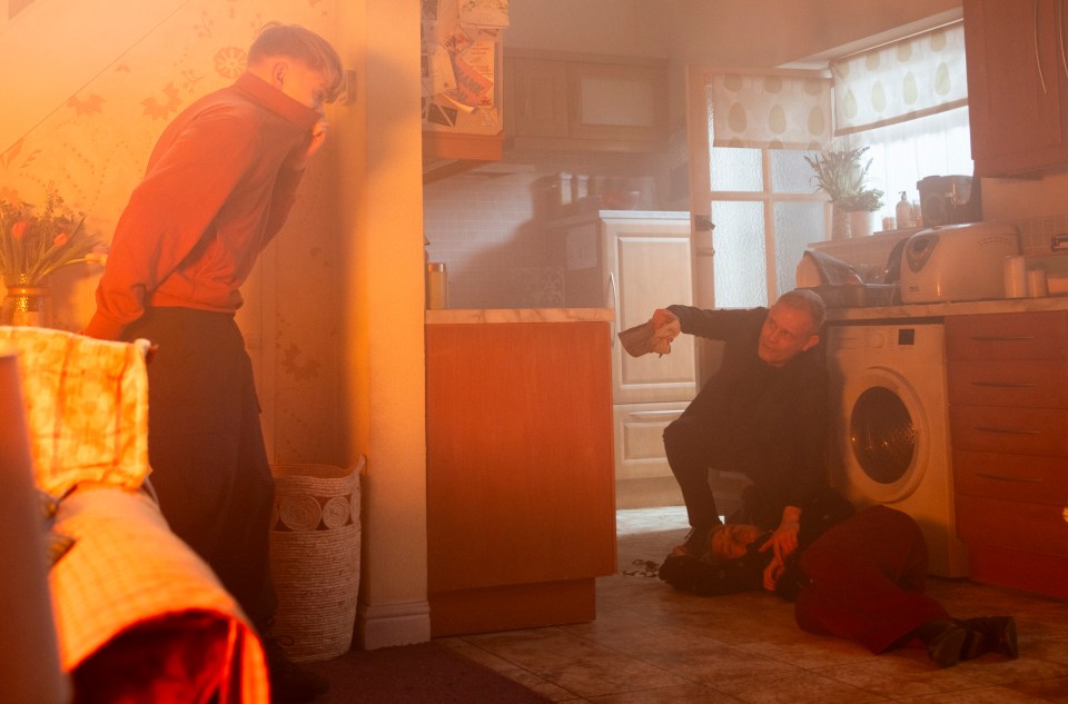 A scene from Coronation Street showing a man kneeling over an unconscious woman on the floor while a boy watches.