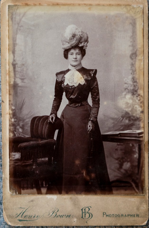 Photo of a woman in a dark dress and large hat, standing next to a chair.