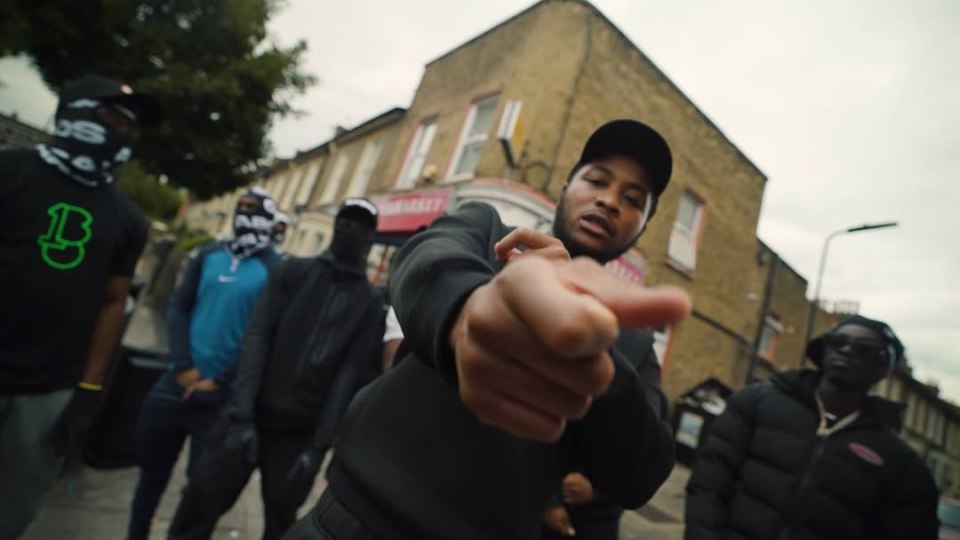 Four gang members on a city street, one in focus pointing towards the camera.