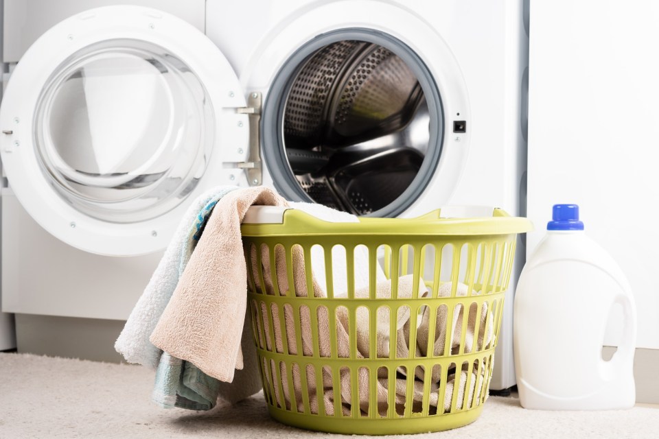 Laundry basket of towels next to a washing machine and detergent.