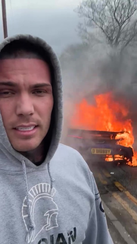 Man in a gray hoodie standing in front of his burning car.