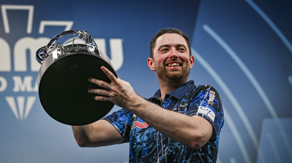 Luke Humphries holding a trophy aloft after winning a darts tournament.