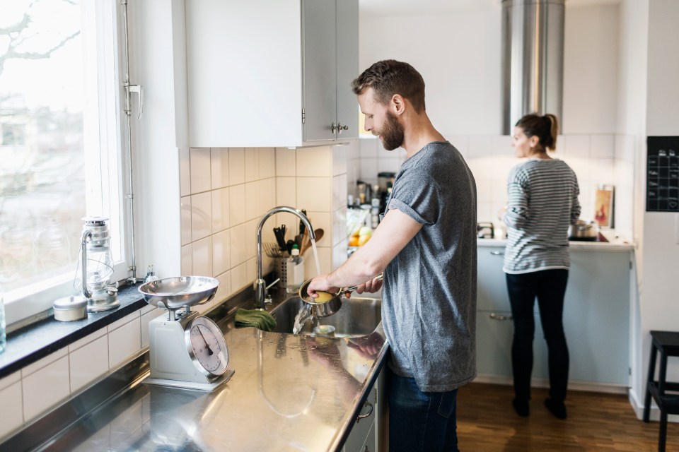 Man washing a saucepan in a kitchen sink, woman standing in the background.