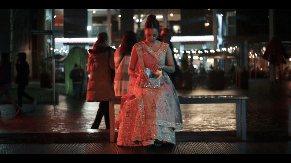 Woman in traditional dress sits on a bench at night, looking at a small purse.