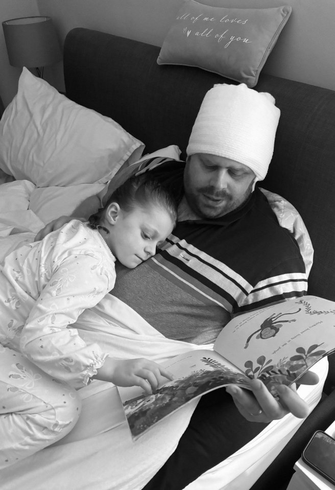 Black and white photo of a father with a head bandage reading a book to his young daughter in bed.