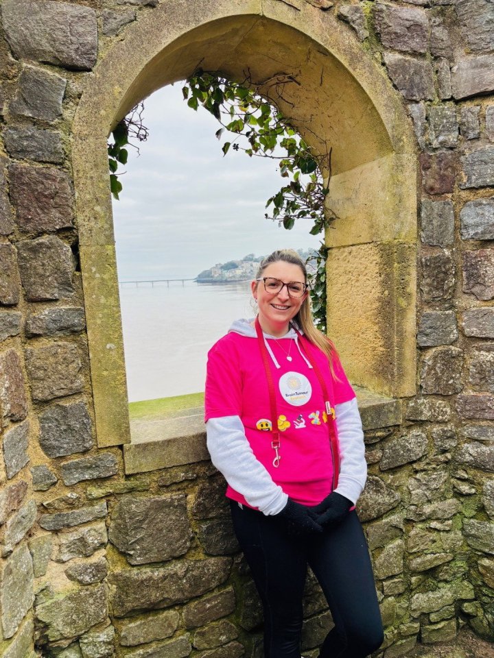 Woman in pink Brain Tumour Research shirt standing in stone archway overlooking water.