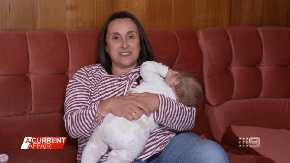 A woman holds her crying baby on a red couch during a television interview.