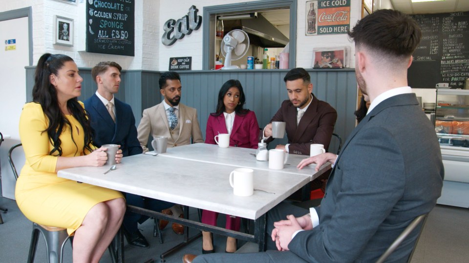 Five contestants from a reality TV show sit around a table in a cafe, talking to a sixth person.