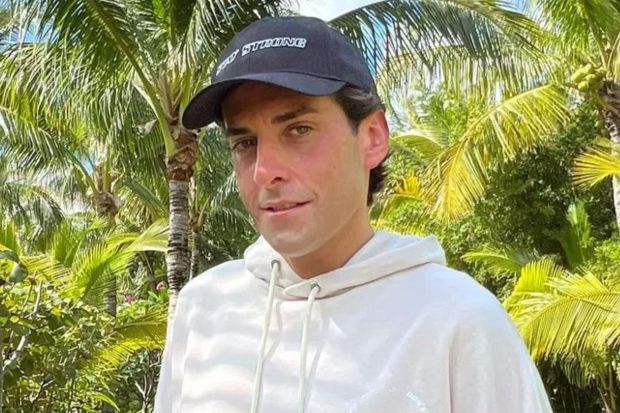 Man wearing a Stay Strong hat and cream hoodie, standing in front of palm trees.