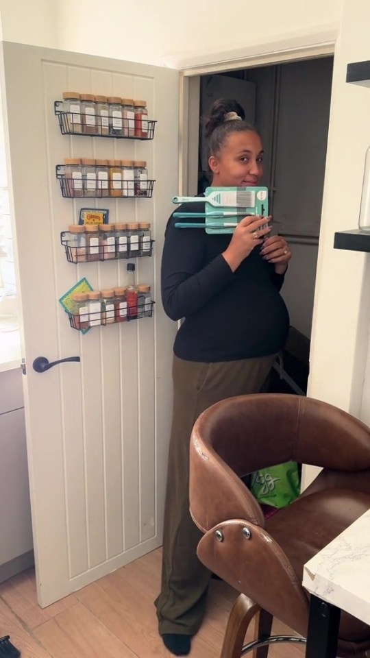 Pregnant woman holding cleaning brushes in front of a kitchen door with spice racks.