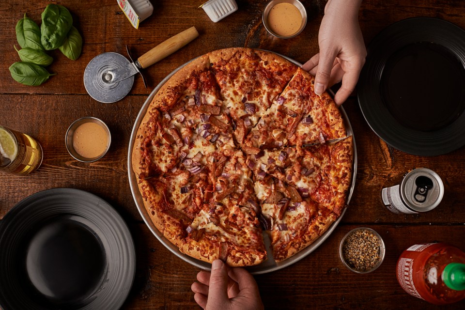 Overhead view of two people reaching for slices of a pizza with red onions and meat.  There are also drinks, dips, and seasonings on the table.