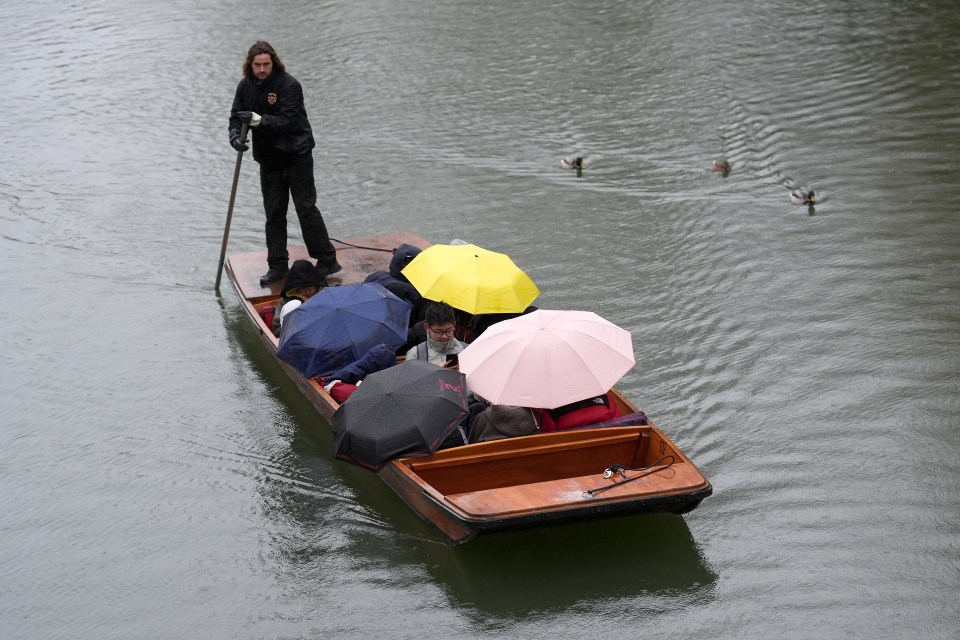 People punting on a river in the rain, using umbrellas for shelter.