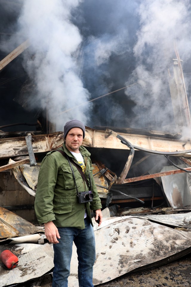 A man stands in front of a smoke-filled, destroyed building.