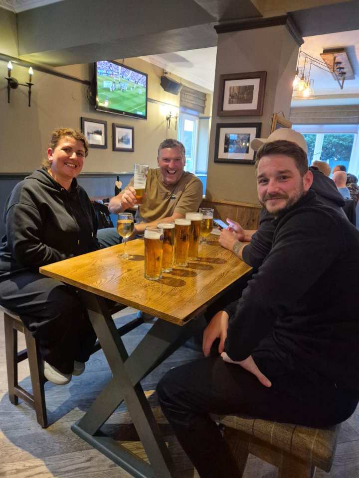 Three people sitting at a pub table with multiple pints of beer.