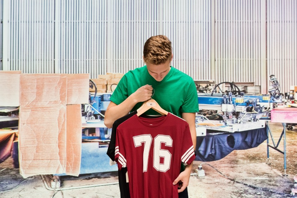 Front view of aged 18-19 years old with brown hair caucasian young male manager at the distribution warehouse in front of merchandise wearing t-shirt who is manufacturing who is working