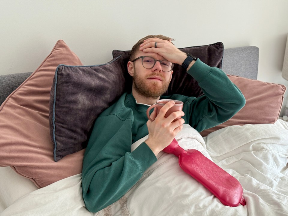 Man in bed holding a mug and hot water bottle, looking unwell.