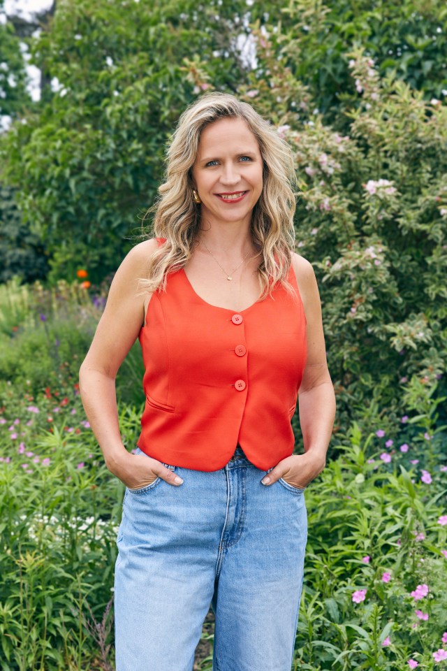 Woman in orange vest and jeans standing in a garden.