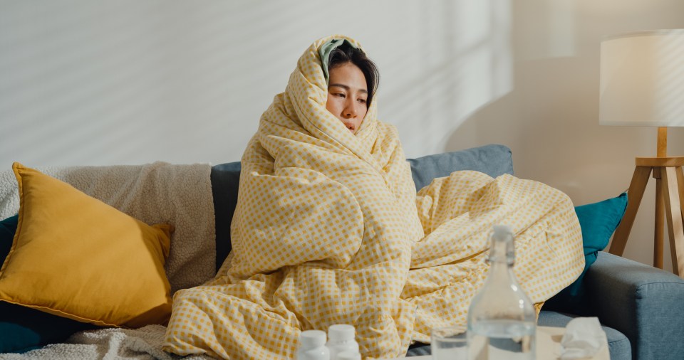 A young woman wrapped in a blanket on a sofa, looking unwell.