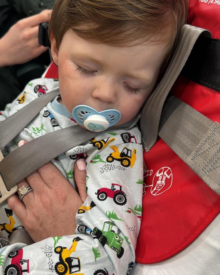 A toddler sleeping in a car seat, wearing pajamas with construction vehicles on them.