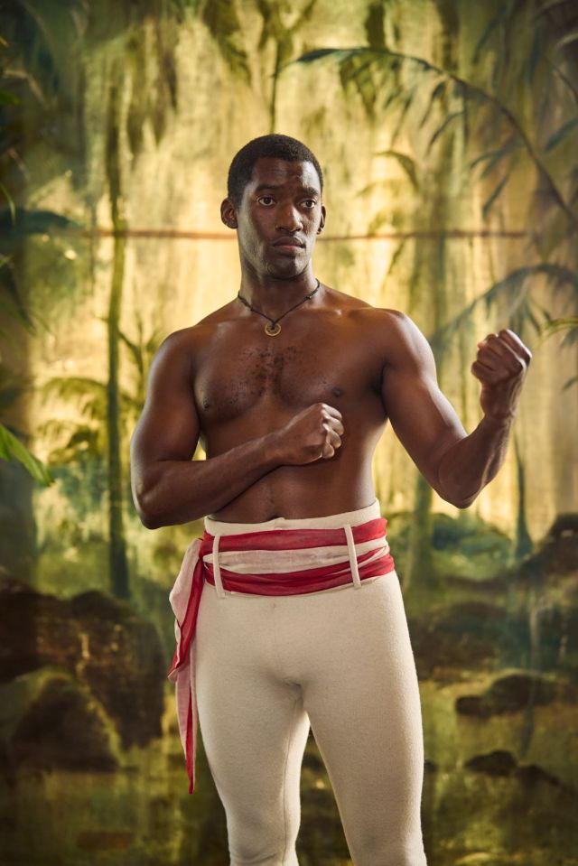 A muscular Black man, shirtless, in boxing stance, wearing cream-colored pants and a red and white sash.