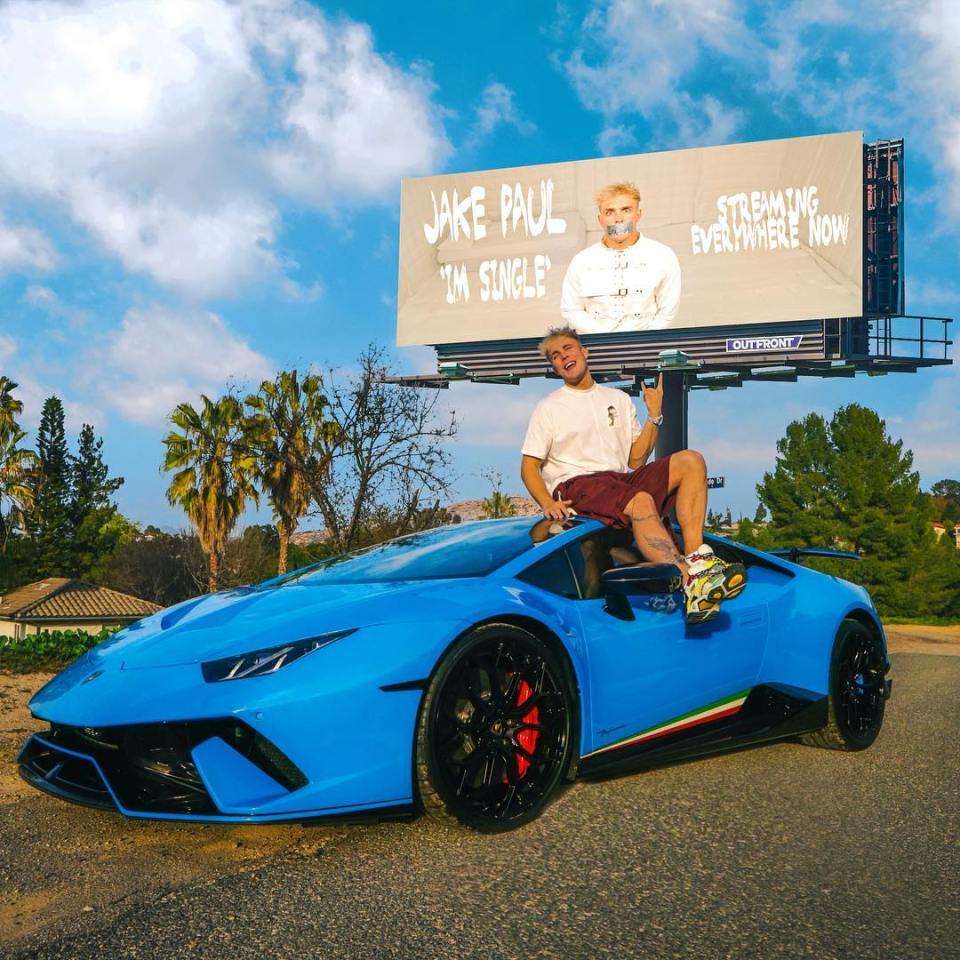 Jake Paul sitting on a blue Lamborghini sports car in front of a billboard advertising his single.