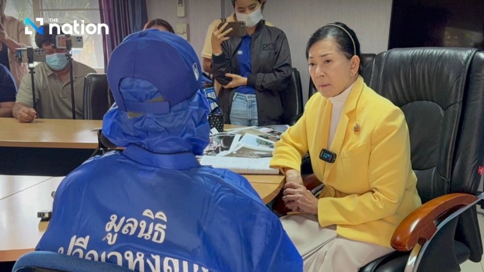 A woman in a yellow jacket sits across from a person in a blue protective suit; several others are in the background.