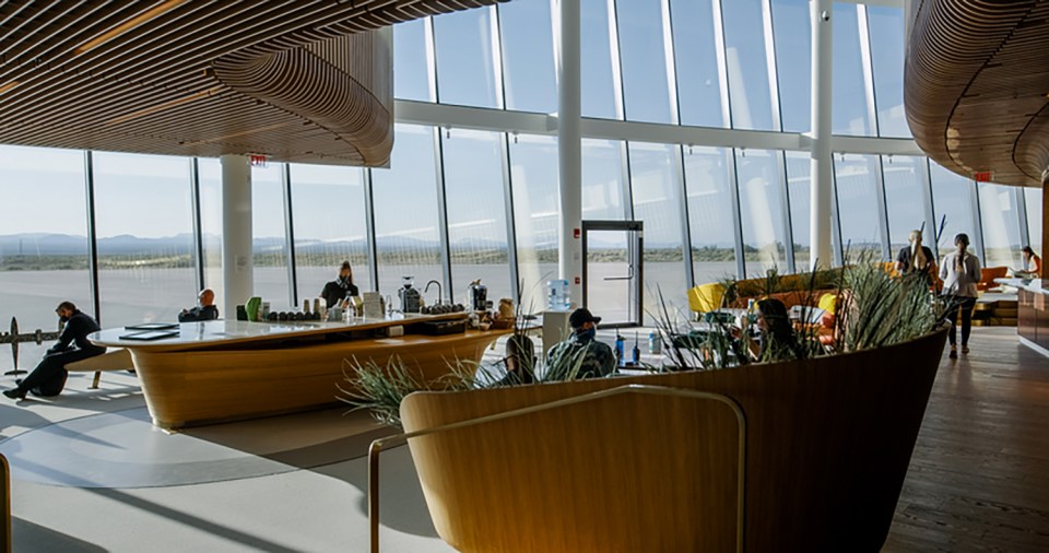 Interior of a modern building with large windows overlooking a landscape, showing people relaxing in a lounge area with a coffee bar.