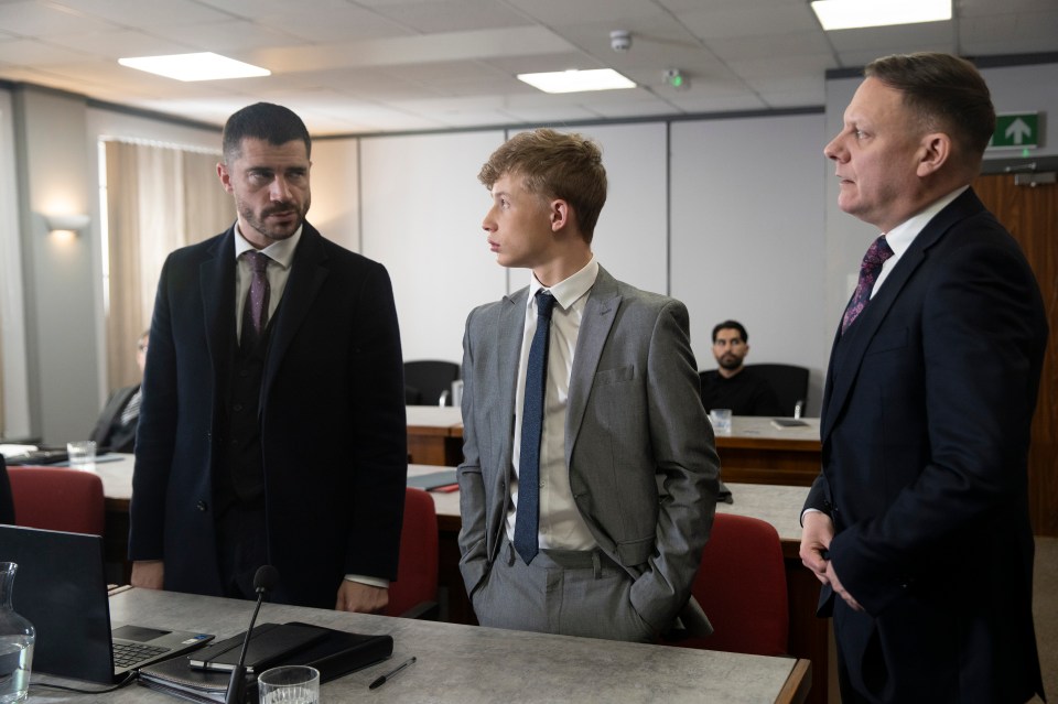 A young man in a grey suit stands between two men in dark suits in a courtroom.