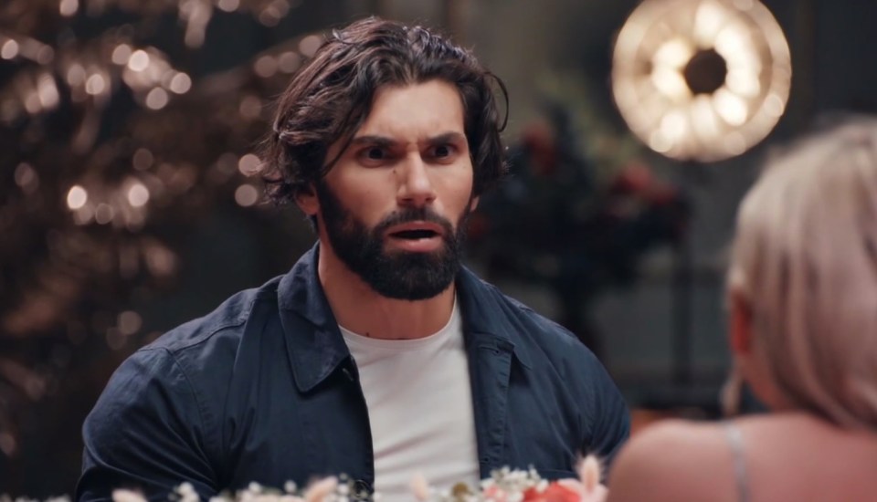 A surprised man with dark hair and a beard, wearing a dark blue shirt, looks at a woman off-camera.