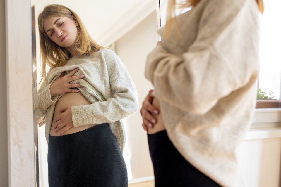 Woman looking in mirror, holding her stomach, experiencing abdominal pain.