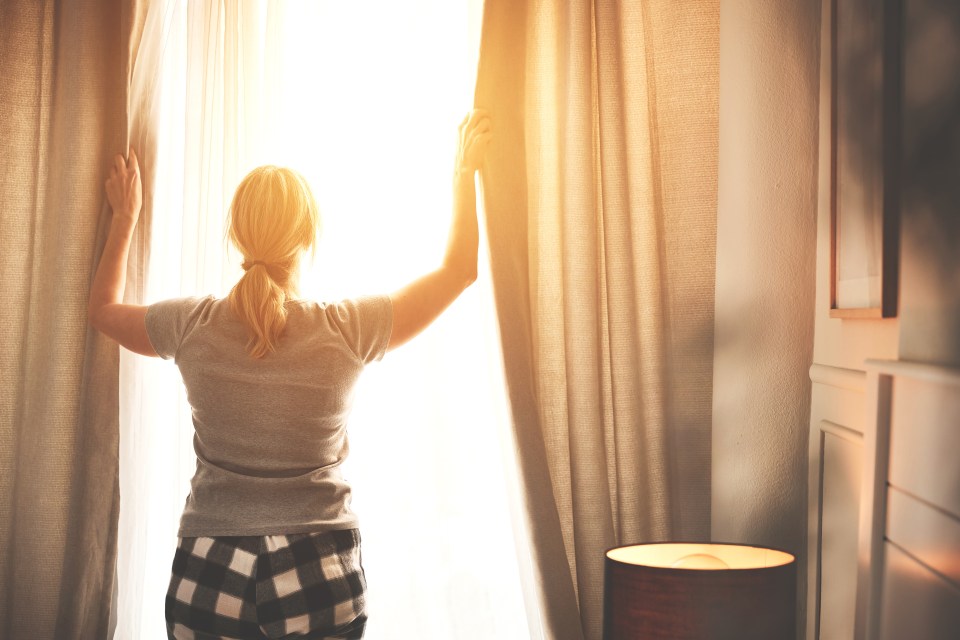 Woman opening curtains in a sunlit bedroom.