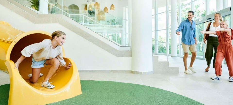 Girl going down a yellow slide in a hotel lobby while a family looks on.