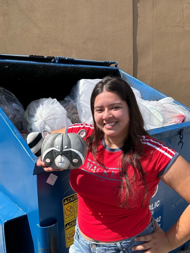 Woman holding a raccoon figurine by a dumpster.