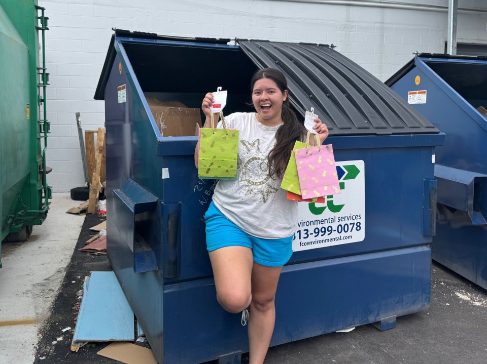 Woman standing by dumpster holding reusable shopping bags filled with salvaged items.