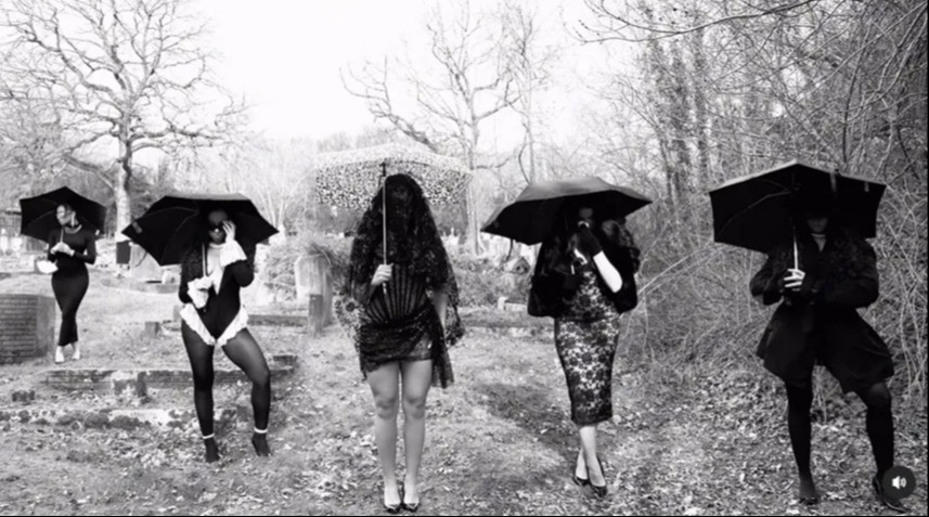 Black and white photo of four women in black dresses and veils, holding umbrellas in a cemetery.