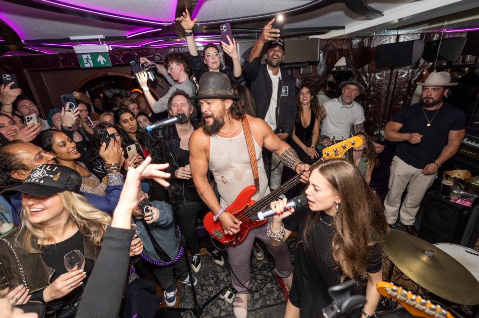 Jason Momoa playing bass guitar on stage with a singer and a crowd of people taking photos.