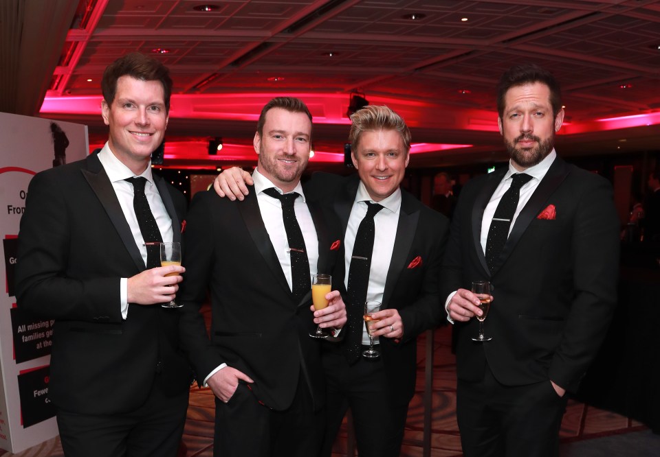 Four men in tuxedos at an event, holding drinks.