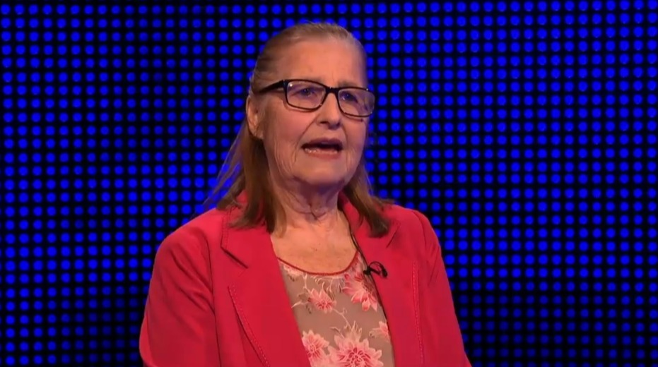 Woman in red jacket and glasses speaking in front of a blue screen.