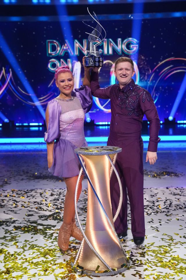 Two ice dancers in purple outfits holding a Dancing on Ice trophy.