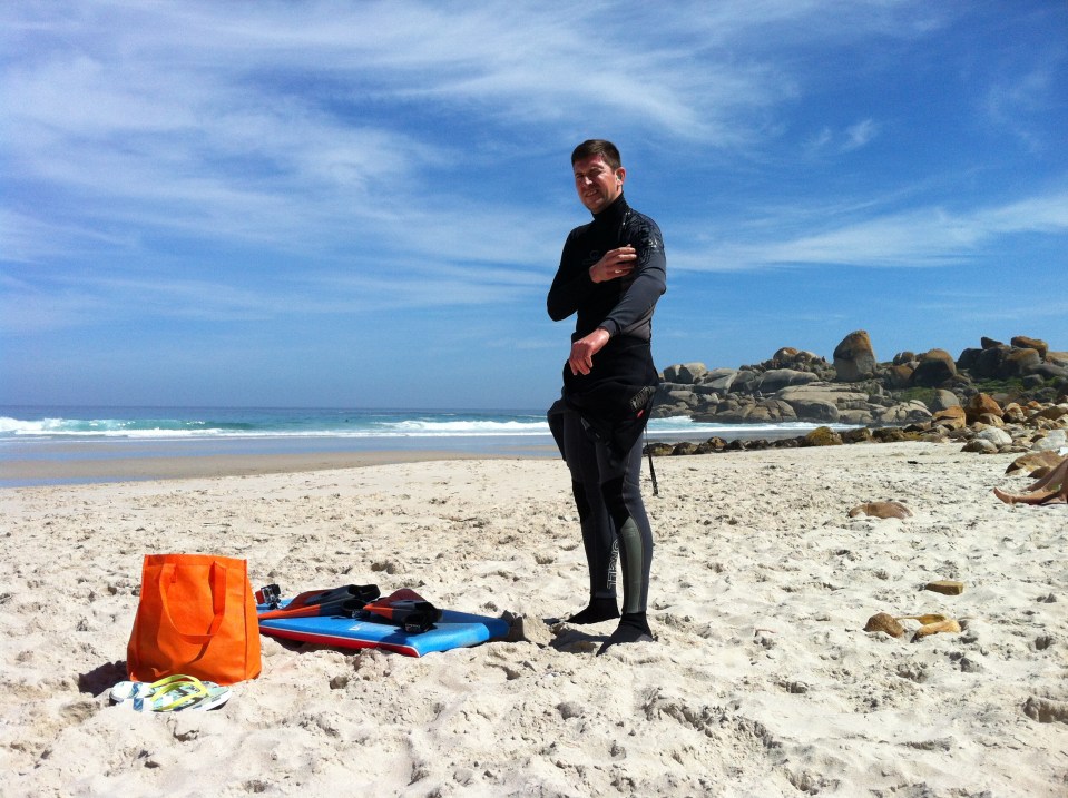 Man in wetsuit on a beach with boogie board and bag.