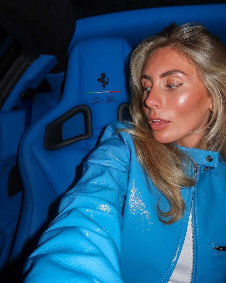 Woman in a blue leather jacket sitting in a blue Ferrari.