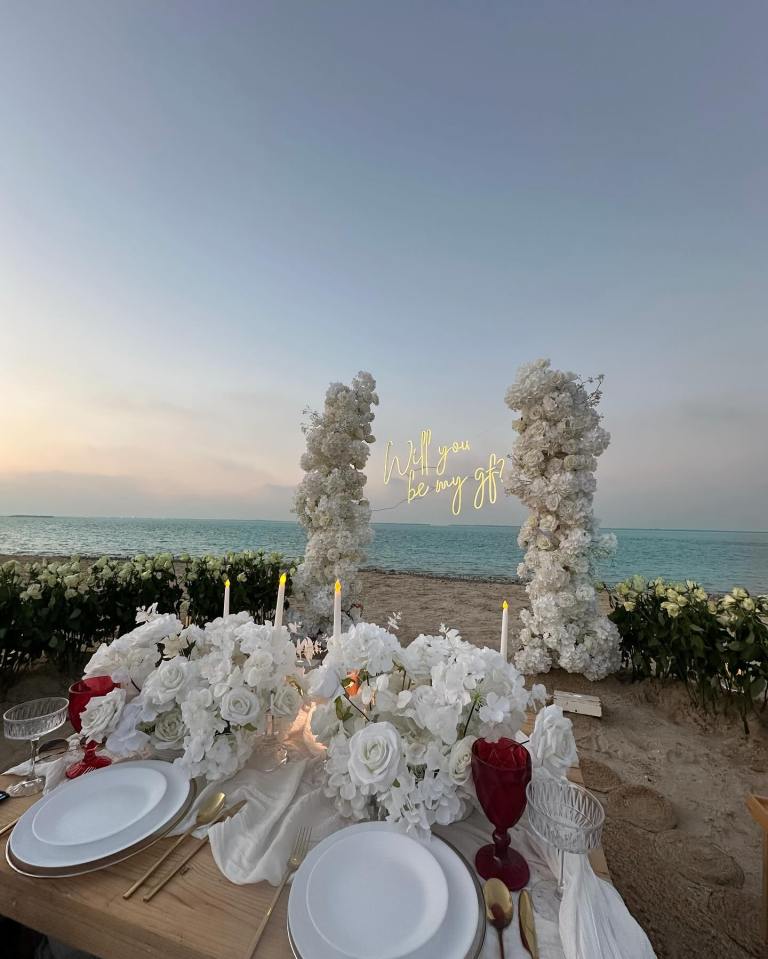 Beach proposal with white flowers and a neon sign asking "Will you be my gf?".