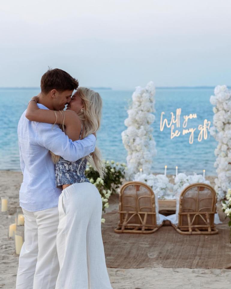 Couple embracing on a beach with a romantic proposal setup in the background.