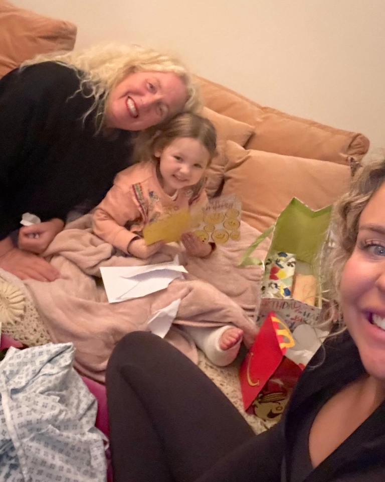 A young girl with a leg cast sits on a couch with her mother and grandmother, surrounded by birthday gifts.